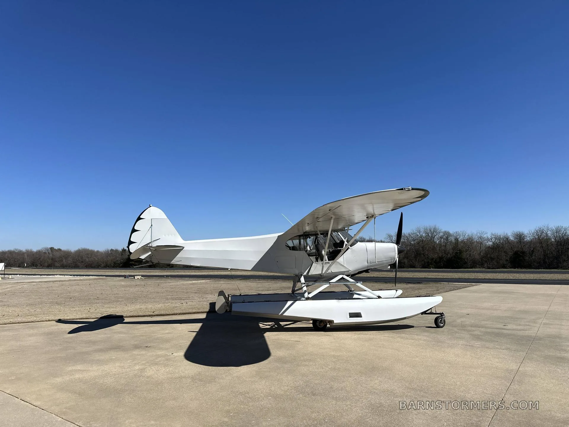 A white seaplane with floats is parked on an asphalt runway under a clear blue sky.