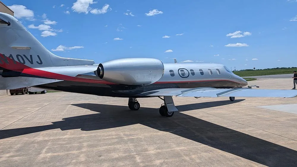 A side view of a silver and black Learjet with a red stripe, showing the tail with registration N10VJ, parked on a tarmac.