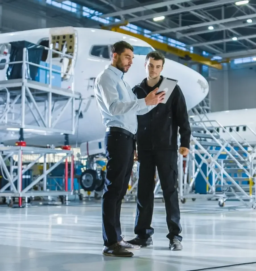 Two men review a tablet in front of a white aircraft undergoing maintenance in a hangar.