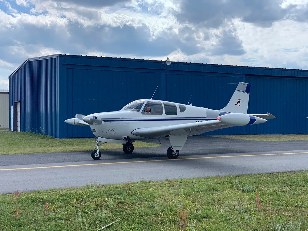 A white and blue Beechcraft Debonair N979Q on a tarmac in front of a blue hangar under a cloudy sky.