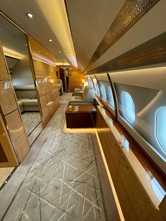 A luxurious aircraft interior features a patterned carpet, wood paneling, and individual seats along a brightly lit aisle.