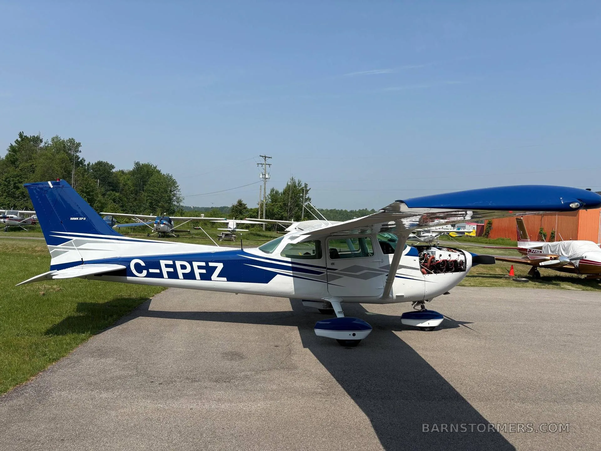 A white and blue Cessna 172 aircraft with registration C-FPFZ is parked on an asphalt ramp with its engine cowling open.