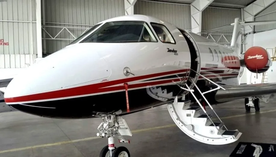The front of a white business jet with red and black stripes is parked inside a hangar with its main door open.
