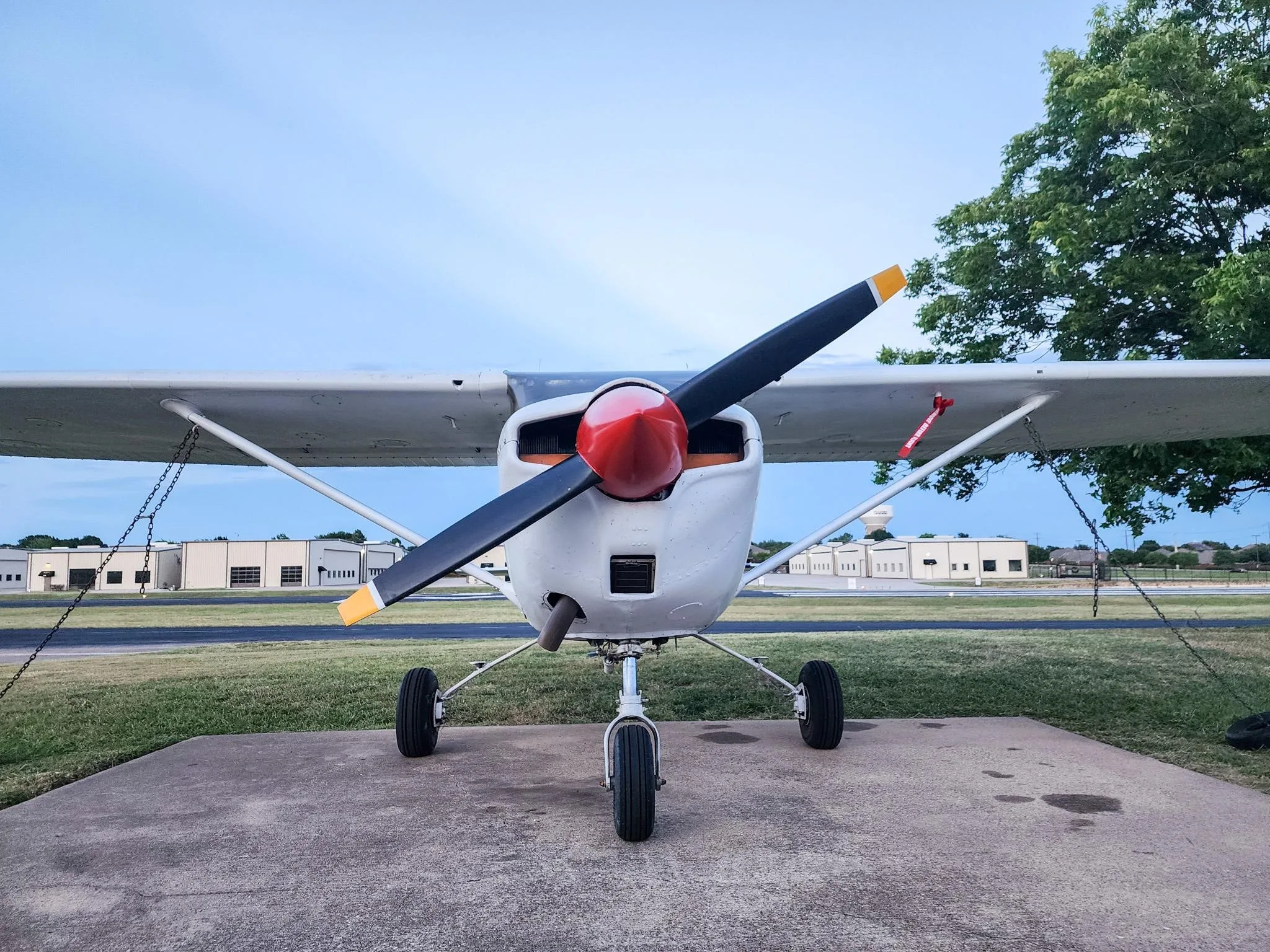 Front view of a white Cessna 150 with a red propeller spinner, parked on a concrete pad at an airfield.