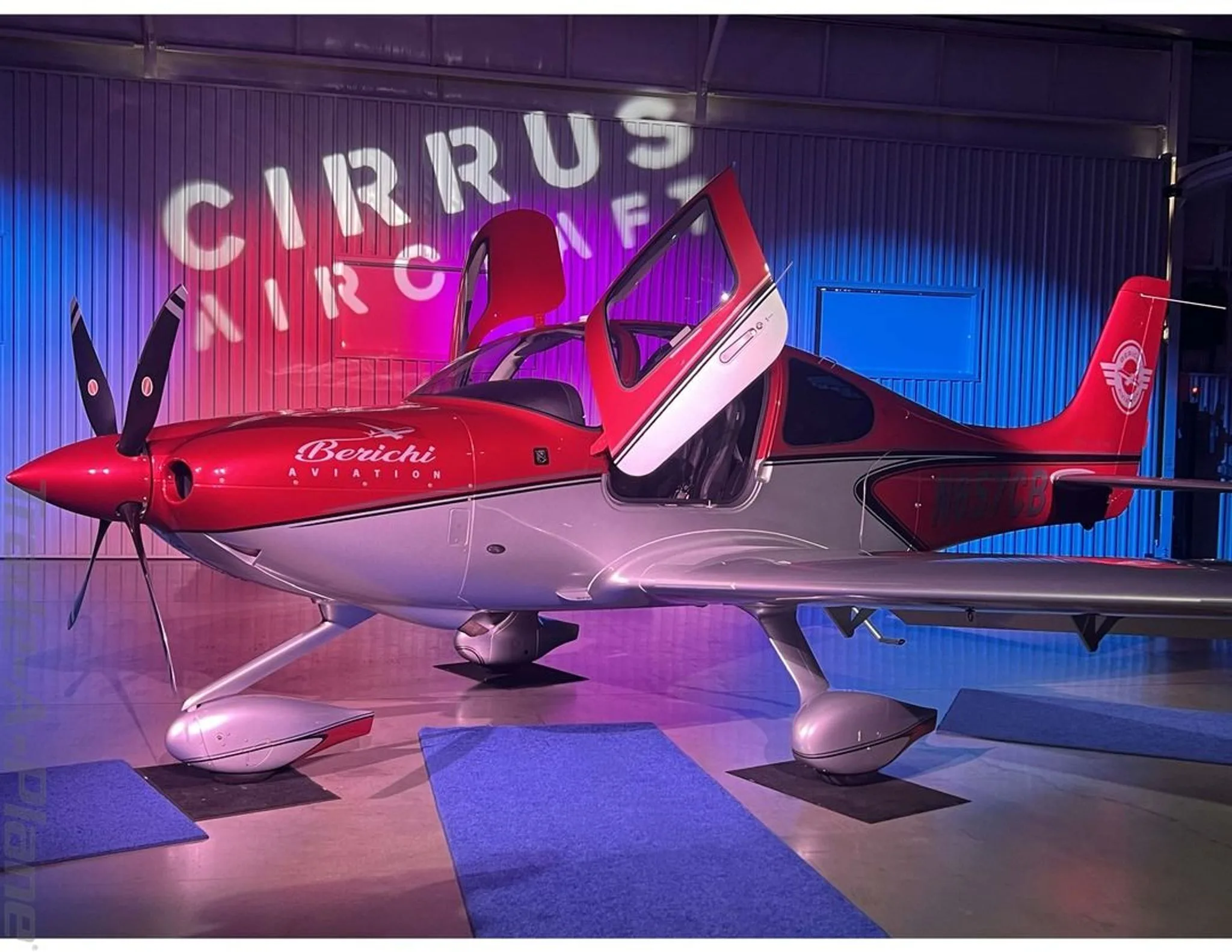 A side view of the red and silver Cirrus SR22 aircraft with its doors open, displayed in a hangar under "Cirrus Aircraft" projection.