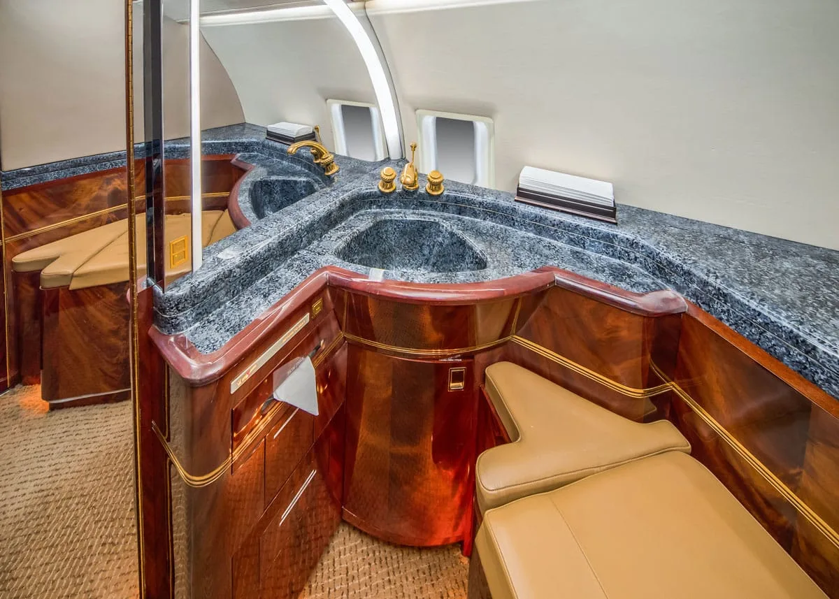 A luxurious aircraft lavatory with dark granite countertops, two sinks, gold fixtures, and rich wooden cabinetry.