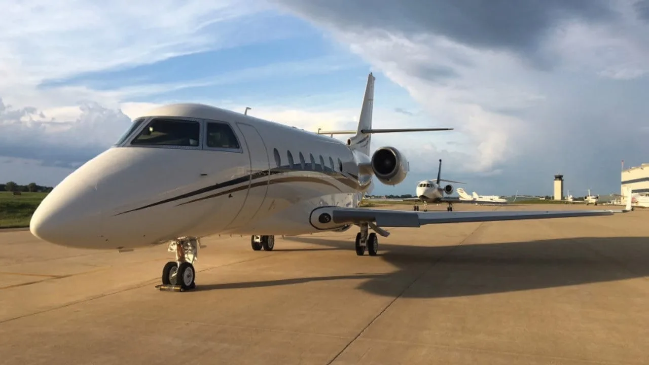 A white business jet with dark and gold stripes parked on an airfield under a partly cloudy sky.