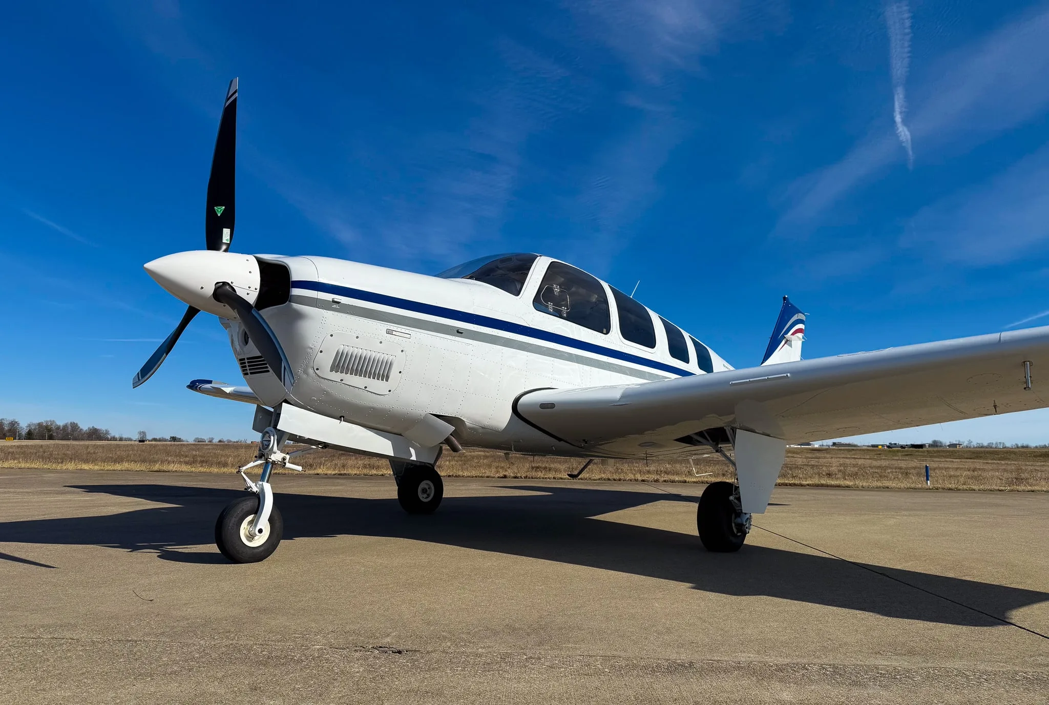 A low-angle front view of a white Beechcraft Bonanza G36 with blue and grey stripes, registration N7CM, parked on an airfield.