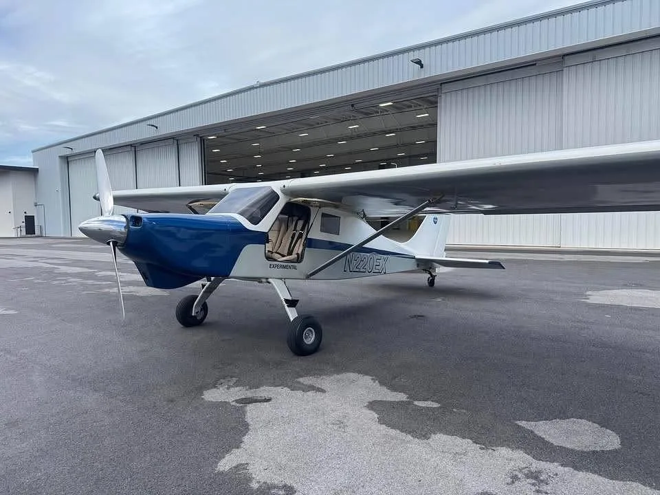 A white and blue experimental aircraft N220EX parked on a tarmac in front of an open hangar.