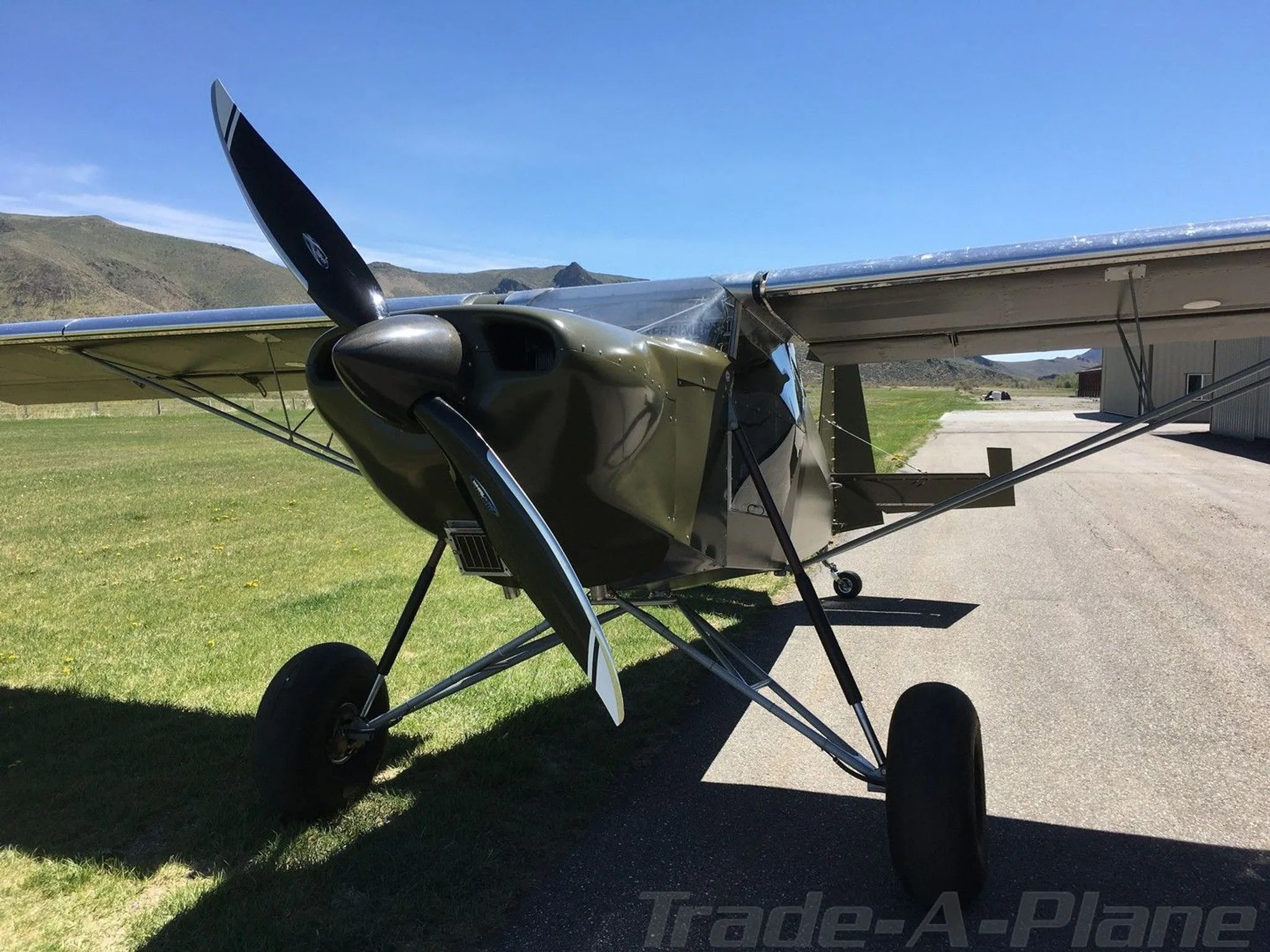 Front view of a green experimental aircraft with large tires parked on an airfield with mountains in the background.