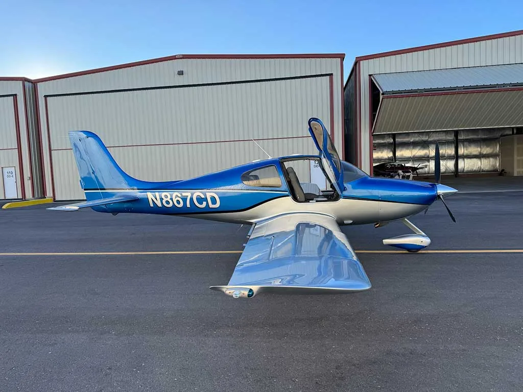 A blue and silver Cirrus SR22 aircraft with registration N867CD, parked on an airfield with its doors open and a hangar door ajar.
