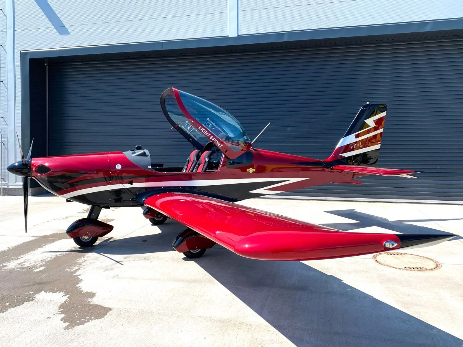 A red and black Viper SD4 light sport aircraft with its canopy open, parked on a concrete apron in front of a hangar.