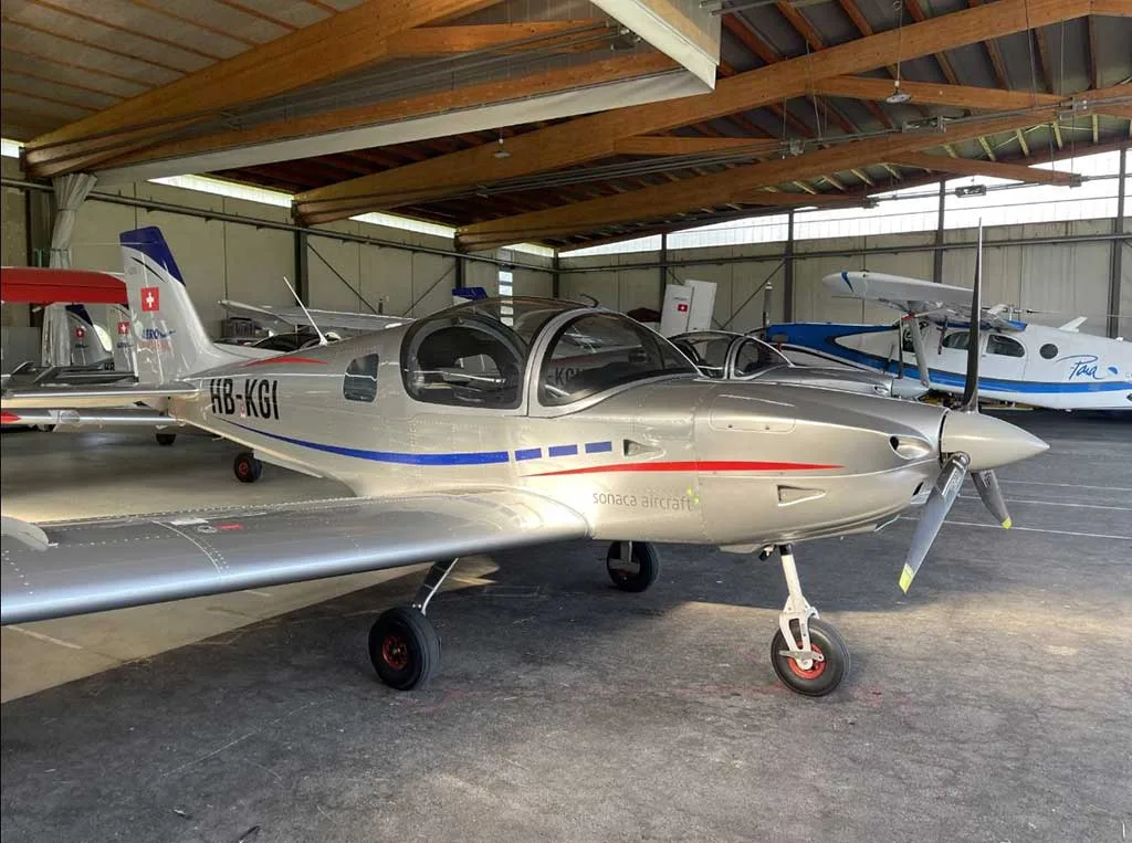 A silver Sonaca 200 aircraft with blue and red stripes, registration HB-KGI, parked inside a hangar.