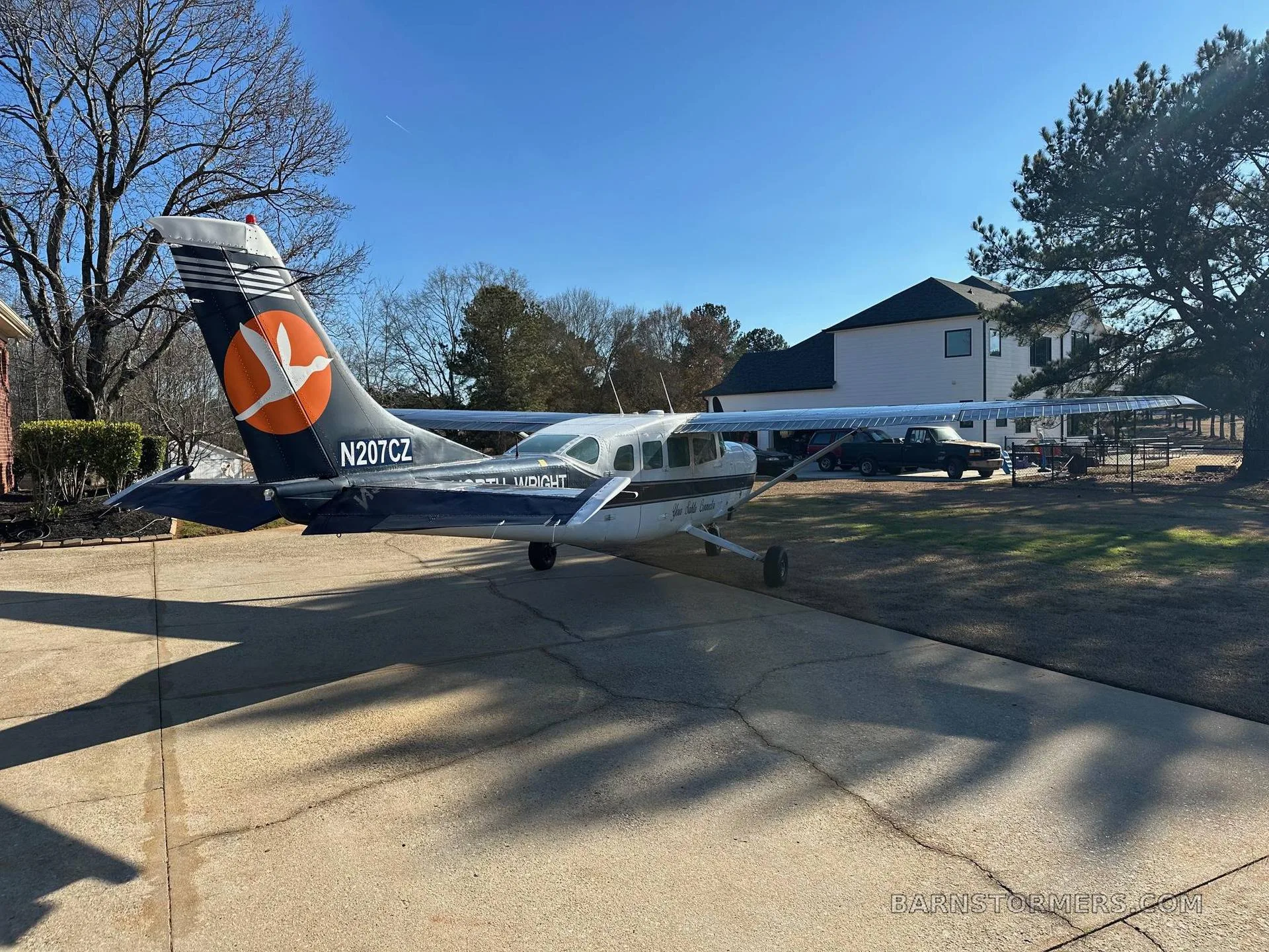 A white and dark blue Cessna 207 aircraft, N207CZ, with an orange bird logo on its tail, parked on a concrete driveway.