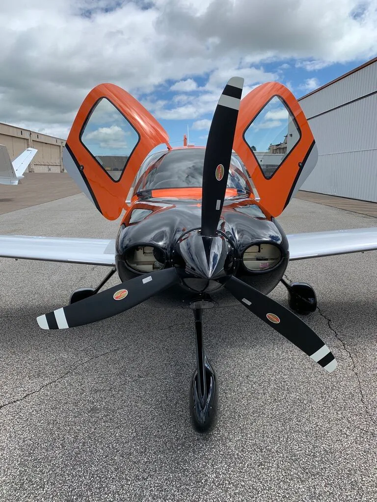 Head-on view of a Cirrus SR20 aircraft with orange, black, and silver paint scheme, doors open, on an airfield.