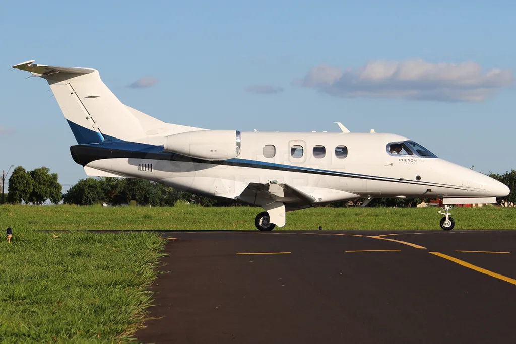 A white Embraer Phenom 100 business jet with blue and black stripes is taxiing on an airport runway under a partly cloudy sky.