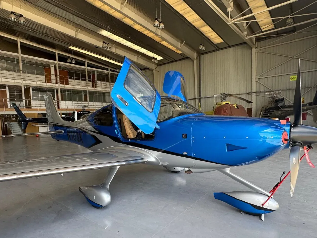 A blue and silver Cirrus SR22 aircraft with gull-wing doors open, parked inside a hangar.