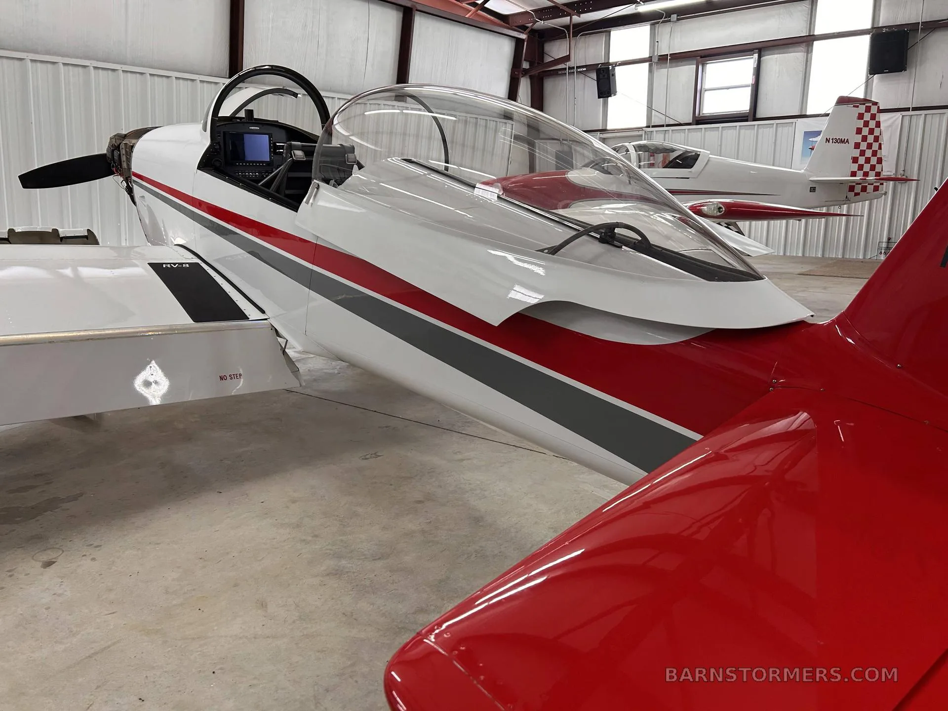 A white aircraft with red and gray stripes and an open canopy, parked inside a hangar with another aircraft in the background.