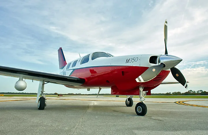 A red and white Piper M350 single-engine turboprop aircraft parked on an airfield under a partly cloudy sky.