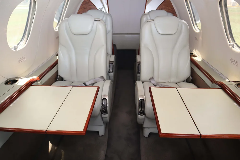 The interior cabin of a private jet, showing light cream leather seats with fold-out tables extended between them.