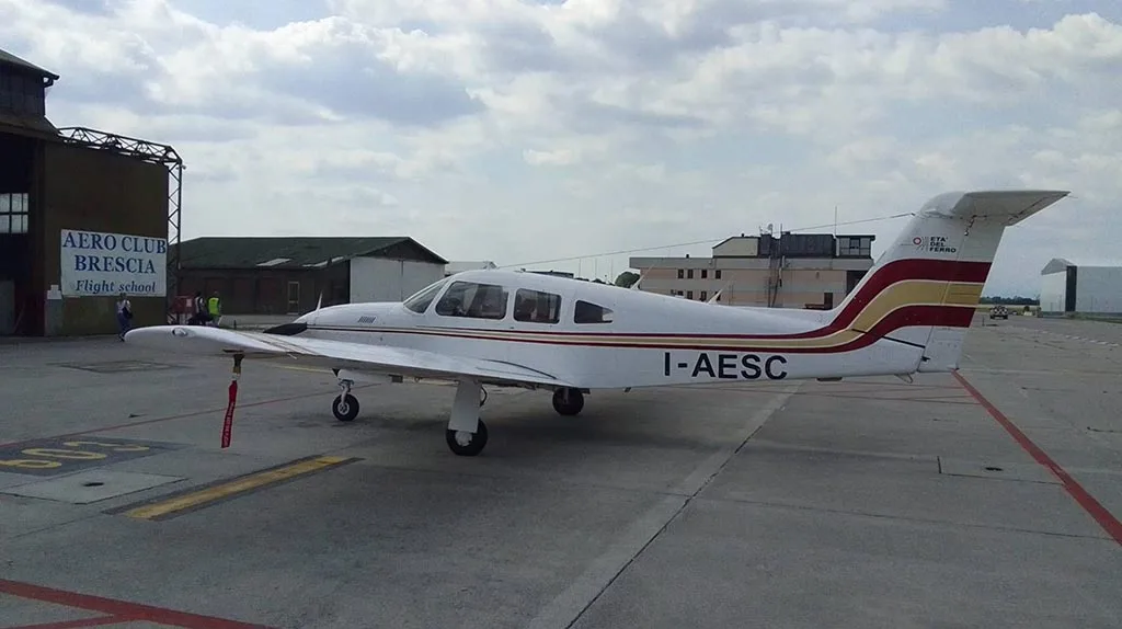 A white Piper PA-28 aircraft with red and gold stripes parked on an airfield next to an "Aero Club Brescia" building.