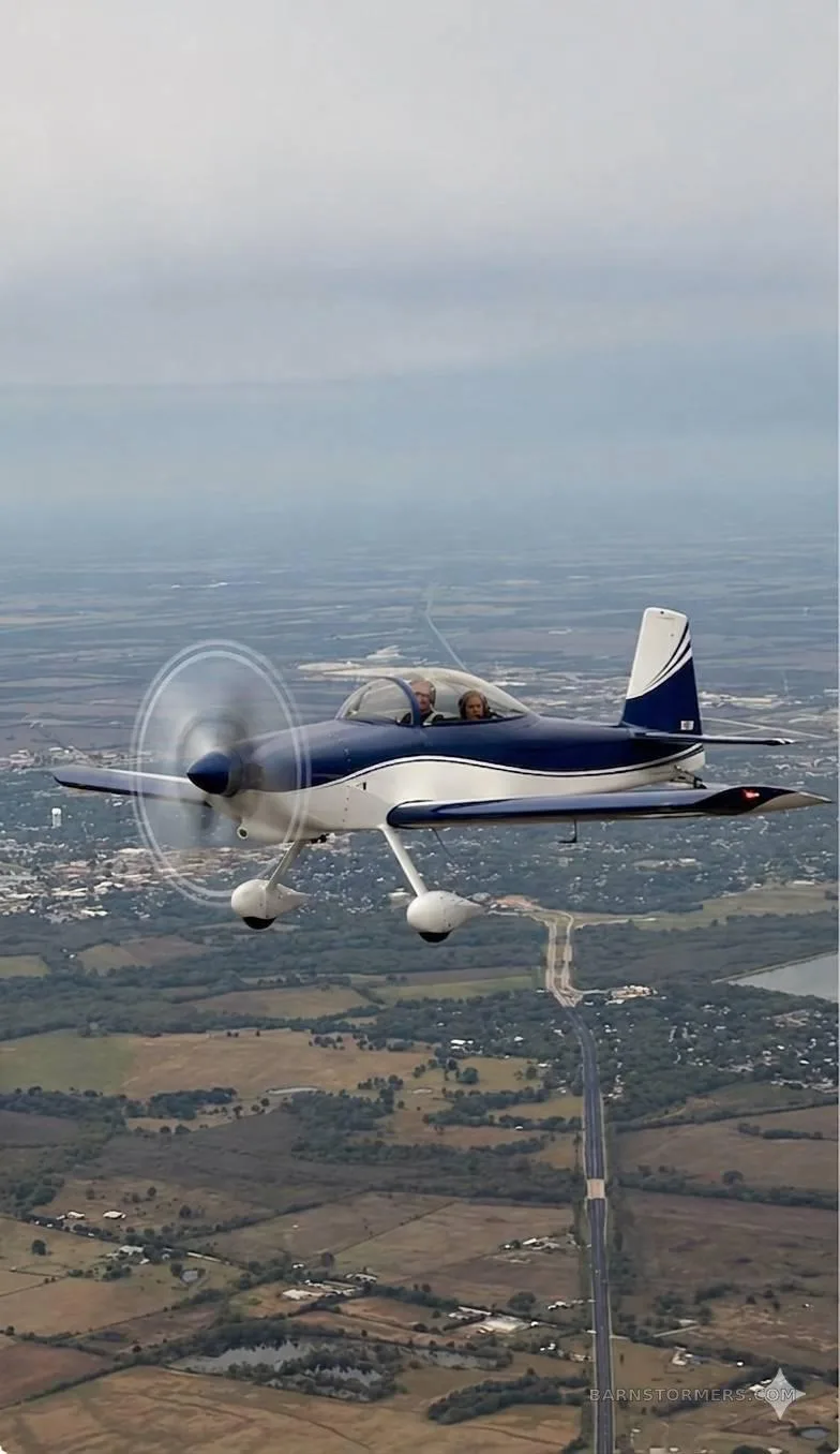 A blue and white single-engine aircraft with two people in the cockpit flies over a rural landscape with a town in the distance.