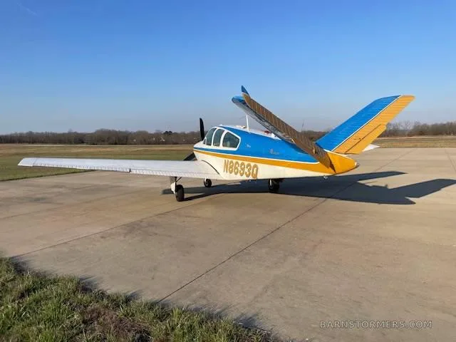 A white, blue, and gold Beechcraft Bonanza with tail number N8693Q is parked on an asphalt ramp, viewed from the rear-left.