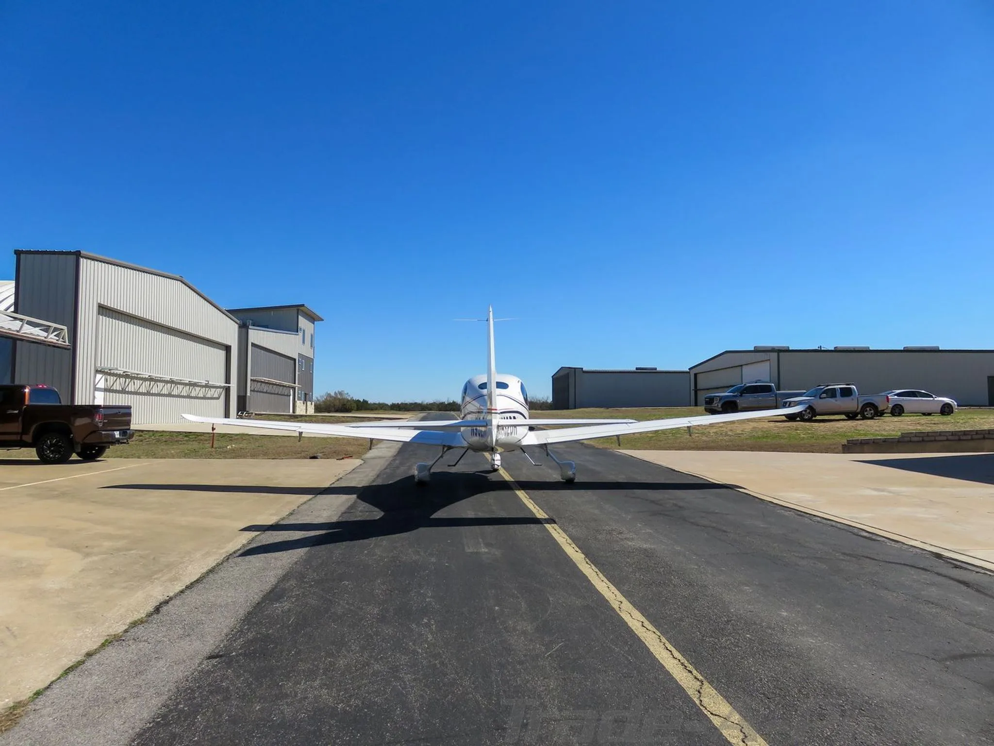 Rear view of a white single-engine aircraft with a single tail, parked on a tarmac with hangars in the background.