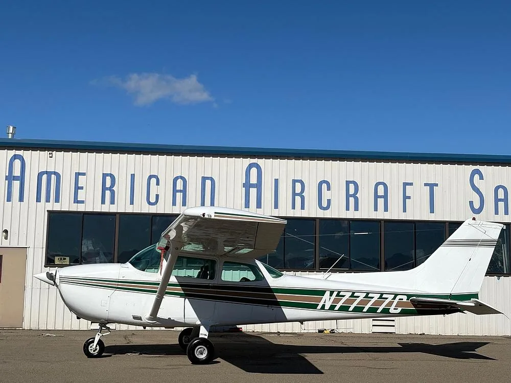 A white Cessna 172 with green and brown stripes, registration N7777C, parked in front of a building.