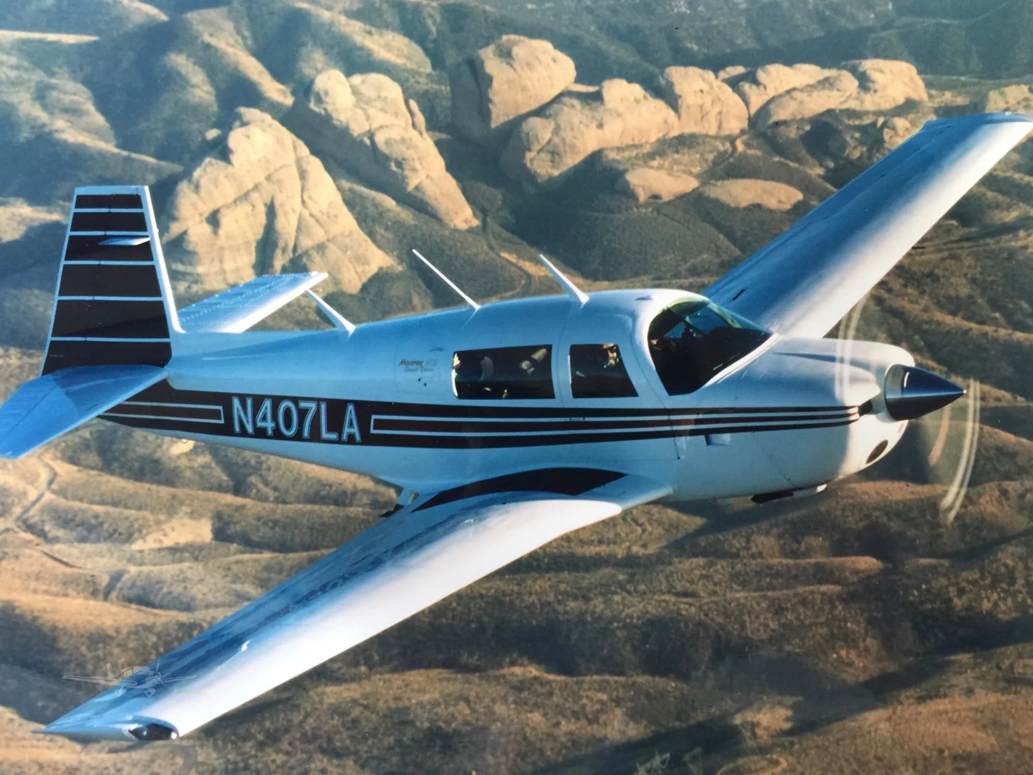 A white Mooney M20 aircraft with dark blue stripes flying over arid, rocky mountains.