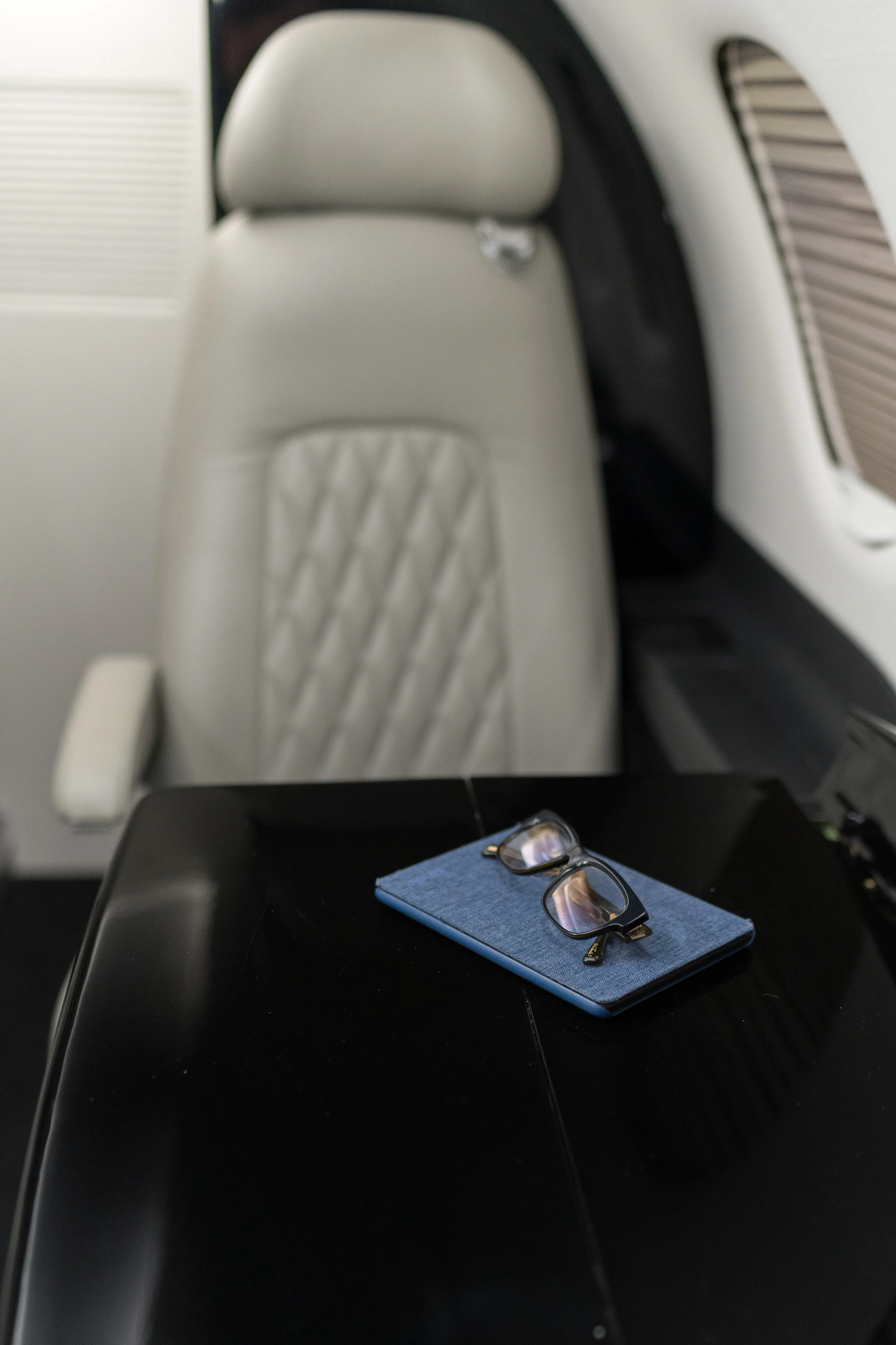 A close-up of a light gray leather cabin seat with a quilted pattern and a black table with eyeglasses and a blue case.