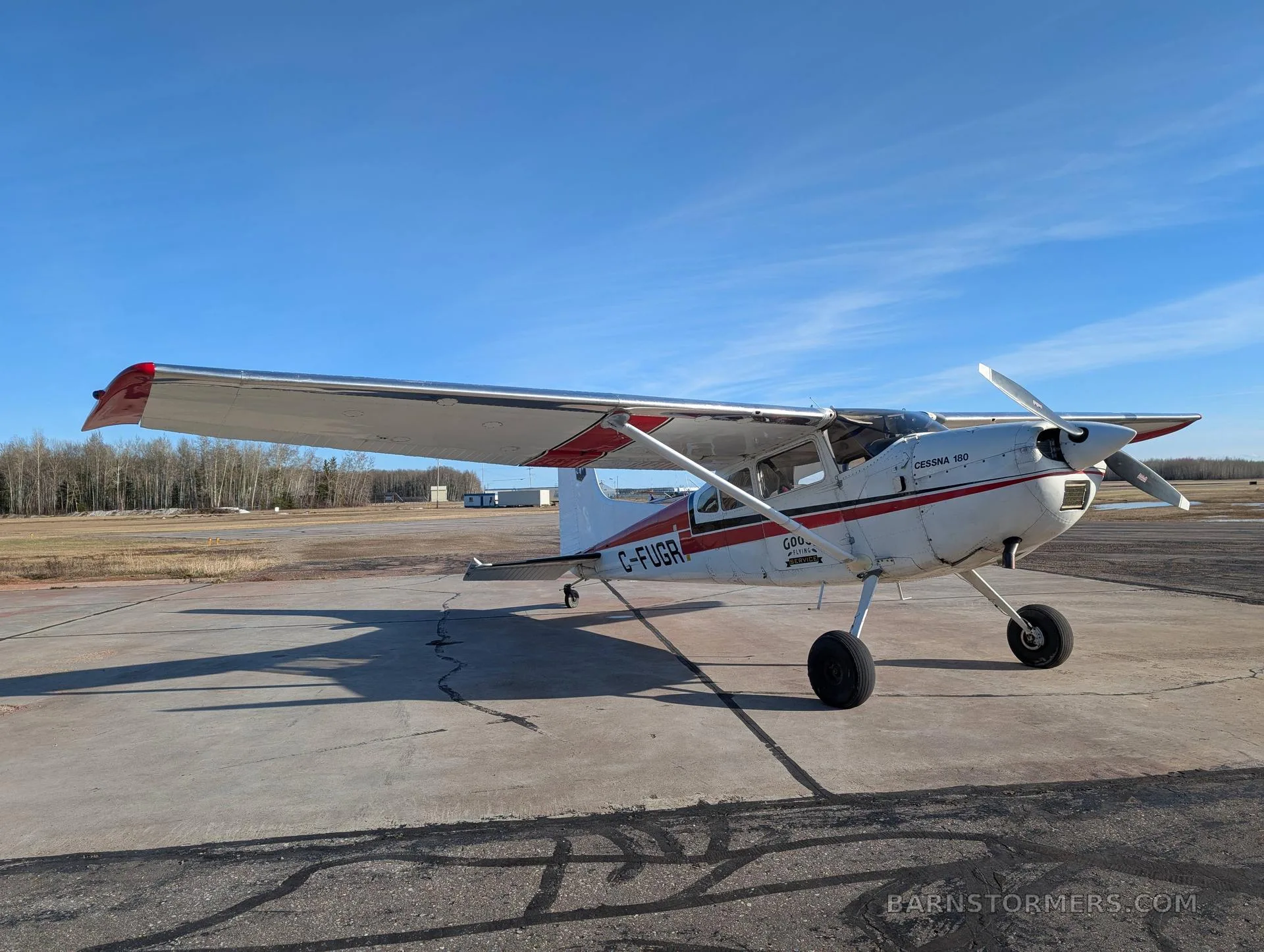 A white and red Cessna 180 (C-FUGR) with a propeller is parked on a concrete tarmac under a clear blue sky.
