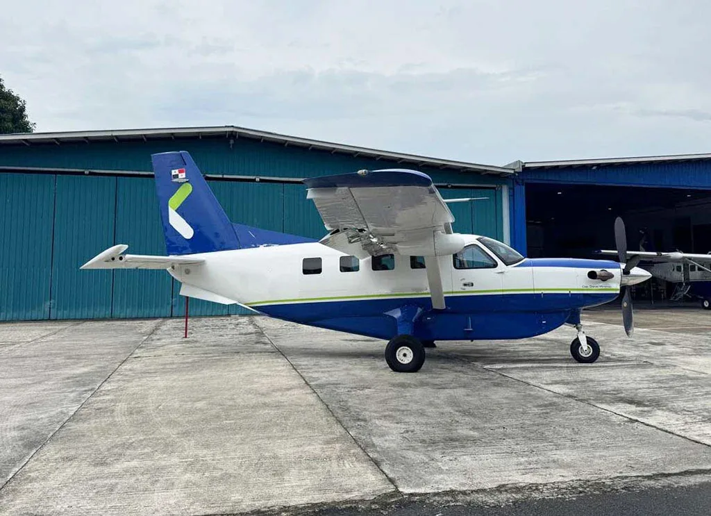 A blue and white Quest Kodiak 100 aircraft with a green tail logo parked on a concrete apron next to a hangar.