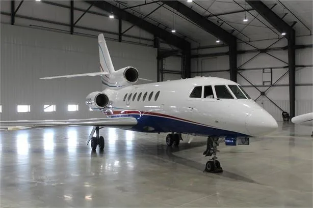 A white and blue Dassault Falcon business jet with red accent stripes is parked inside a large, well-lit hangar.