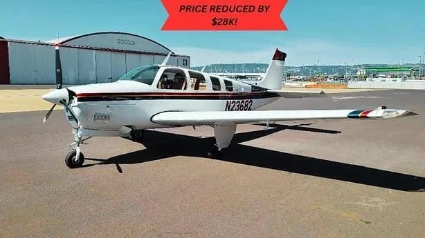 A white Beechcraft Bonanza with a dark stripe and N23682 registration sits on an airport tarmac with a hangar in the background.
