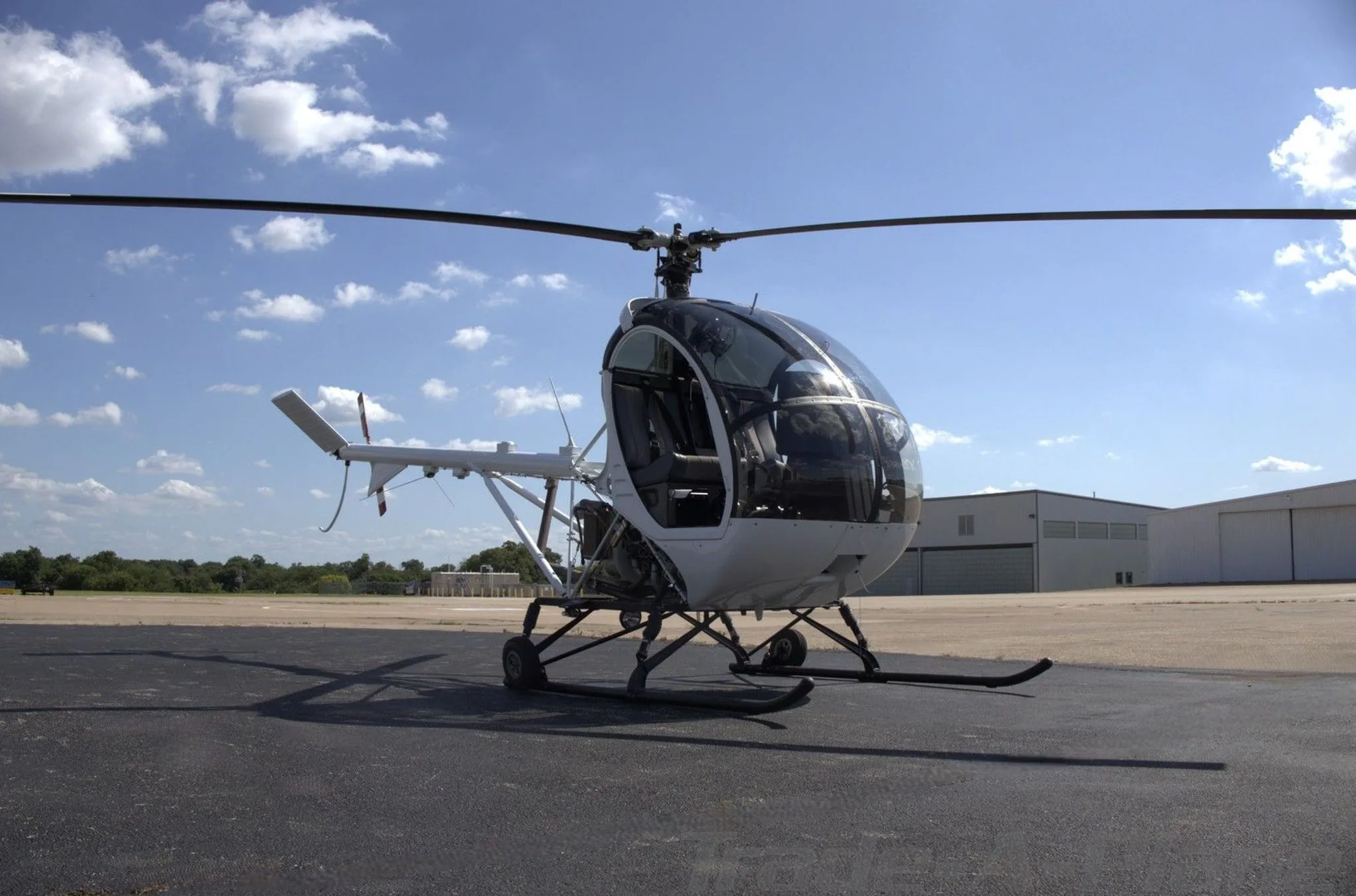 A white Schweizer S300CBi helicopter, registration N2034Z, is parked on an asphalt tarmac with hangars in the background under a partly cloudy sky.