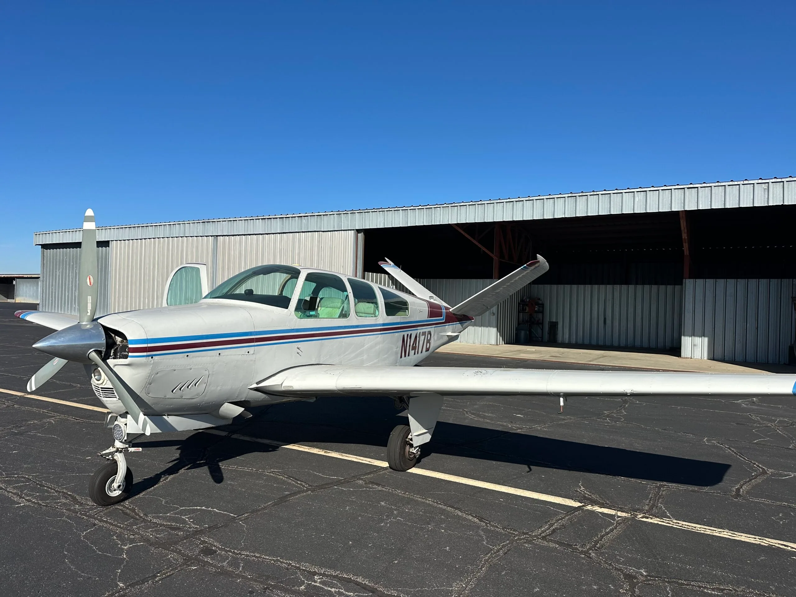 A white Beechcraft Bonanza with blue and maroon stripes is parked on a tarmac in front of an open hangar on a sunny day.