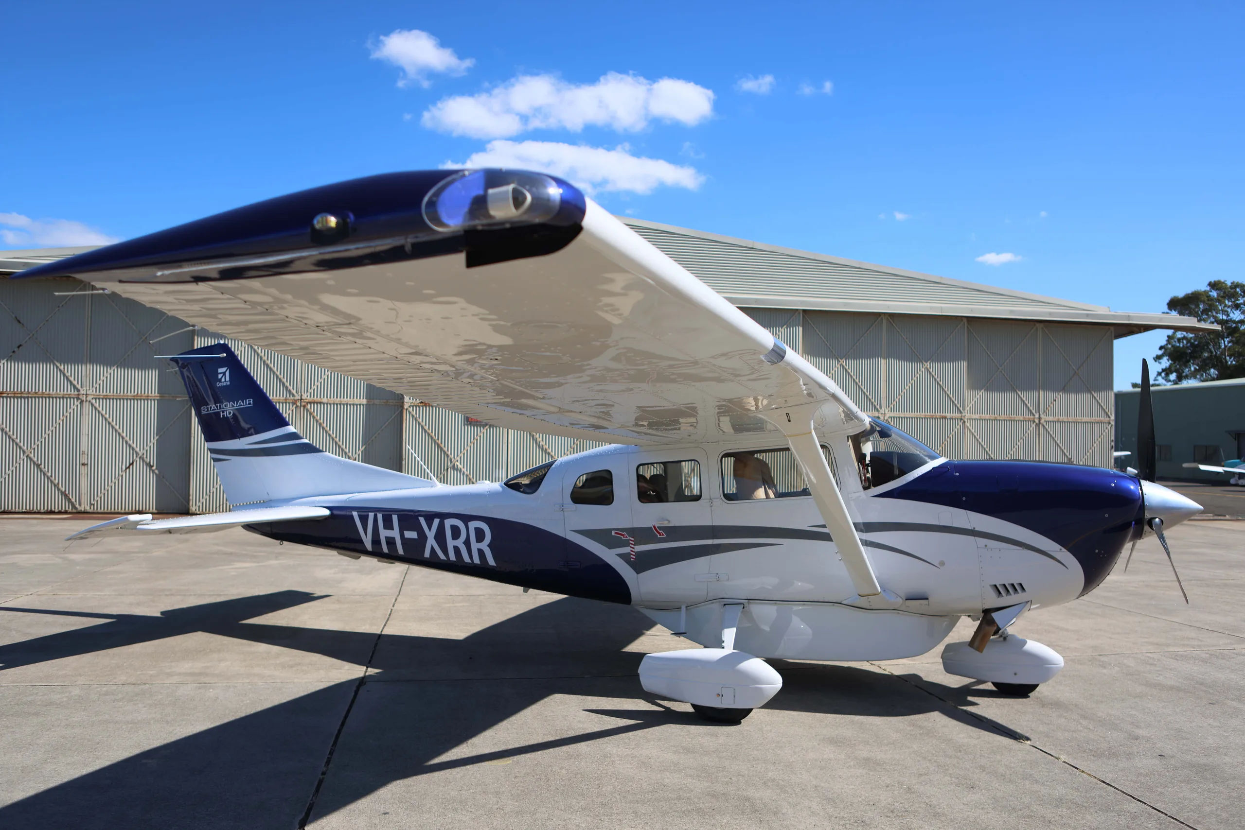 A white and blue Cessna 206 Stationair HD aircraft with registration VH-XRR is parked on an airfield in front of a hangar under a blue sky.