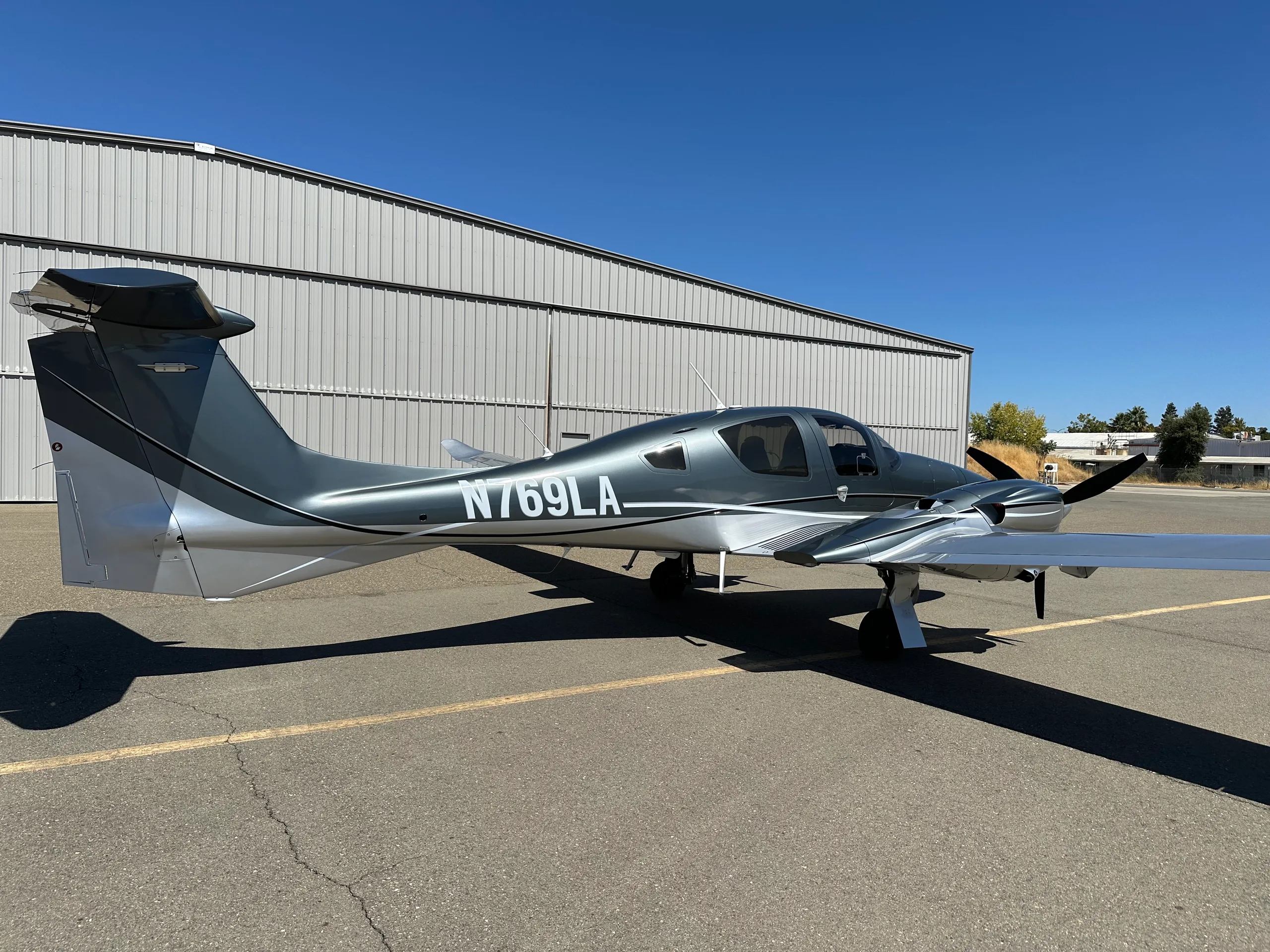 A rear-quarter view of a grey and silver Diamond DA62 aircraft with registration N769LA parked on an airfield.