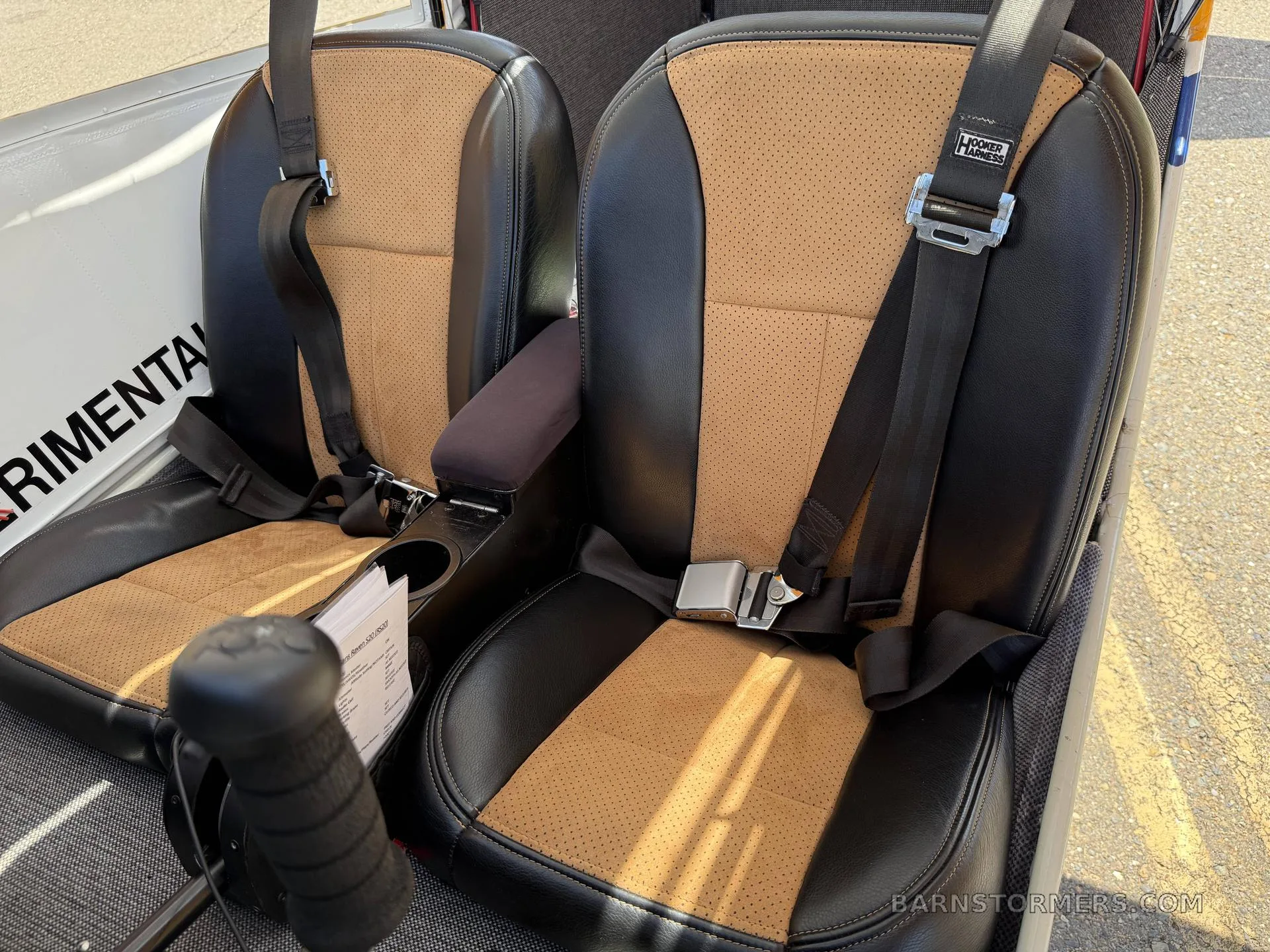 Two black and tan perforated leather aircraft seats with seatbelts and a control stick in the foreground.