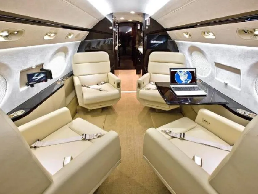 The luxurious interior cabin of a private jet with cream leather seats and a laptop on a table.