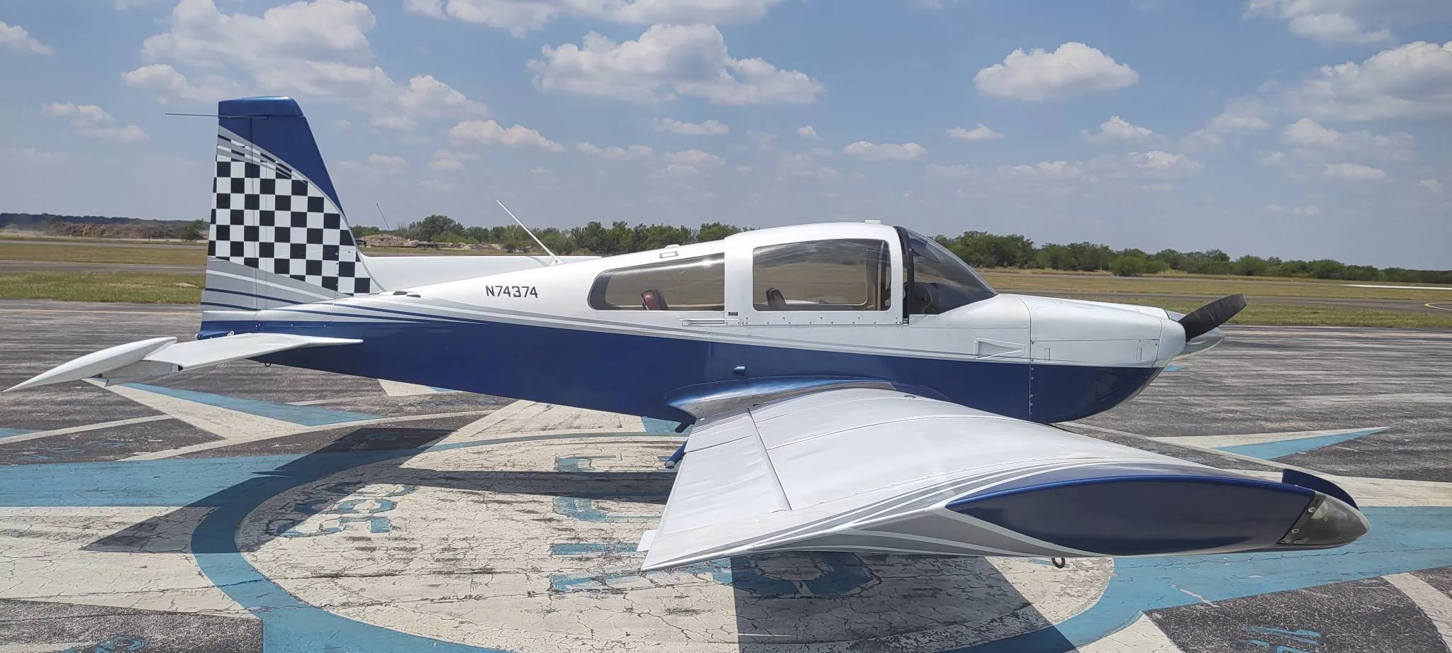 A blue and white single-engine aircraft, N74374, with a checkered tail, parked on an airfield compass rose.