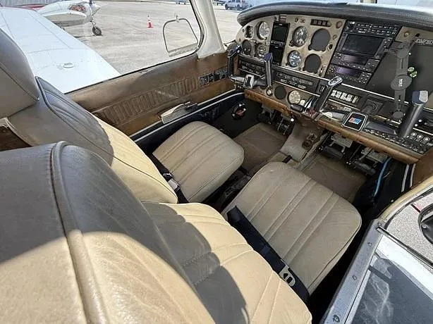 The light brown leather front seats of a small aircraft cabin, with a partial view of the cockpit and an airfield outside.