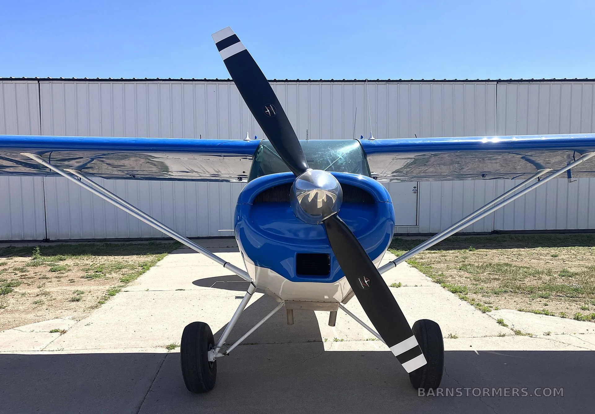 A head-on view of a blue and white high-wing aircraft with a two-bladed propeller, parked on an apron.