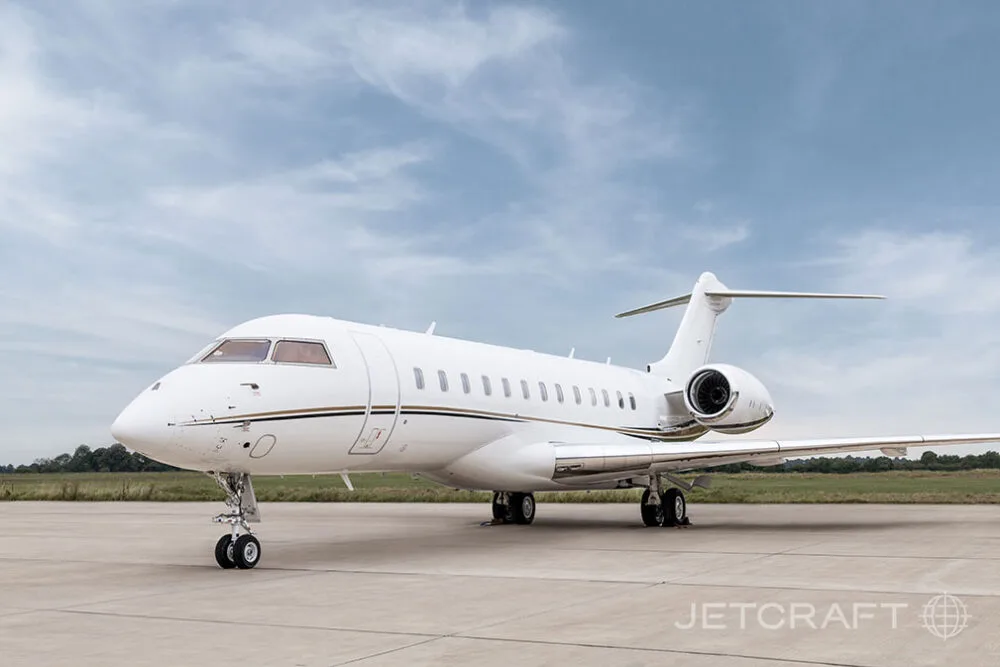 A white Bombardier Global business jet with gold and black stripes parked on an airfield, viewed from the front.