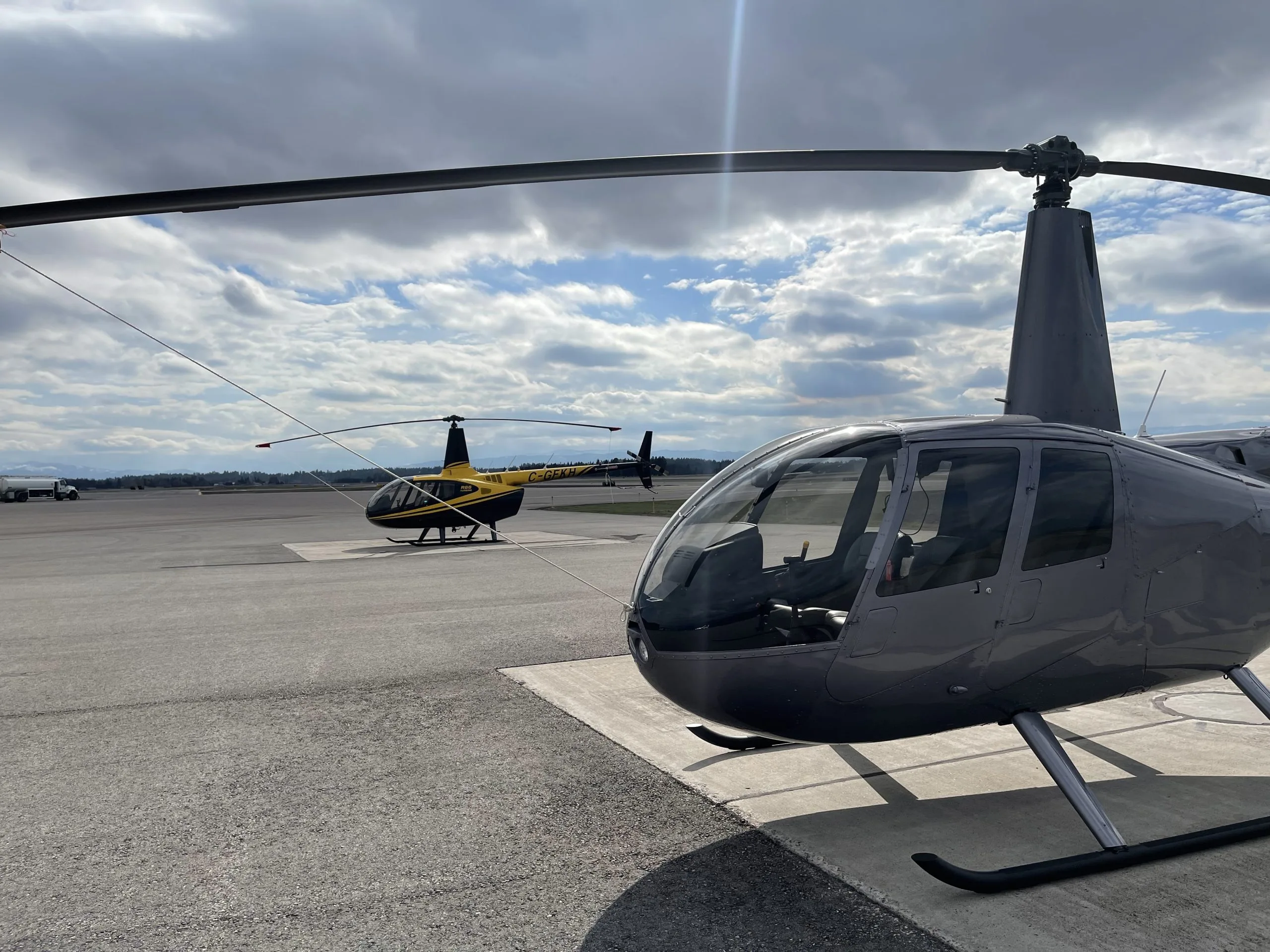 Two Robinson R44 helicopters, one grey in the foreground and one yellow in the background, parked on an airport tarmac under a cloudy sky.