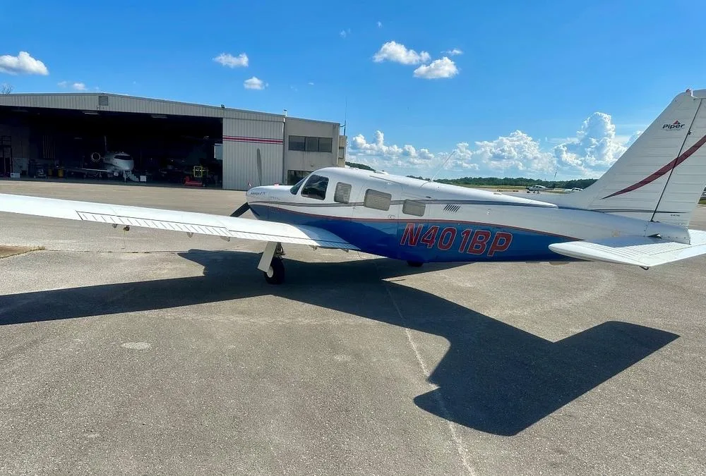 A Piper Saratoga II TC aircraft, N401BP, in white and blue, parked on an airport ramp with a hangar in the background.