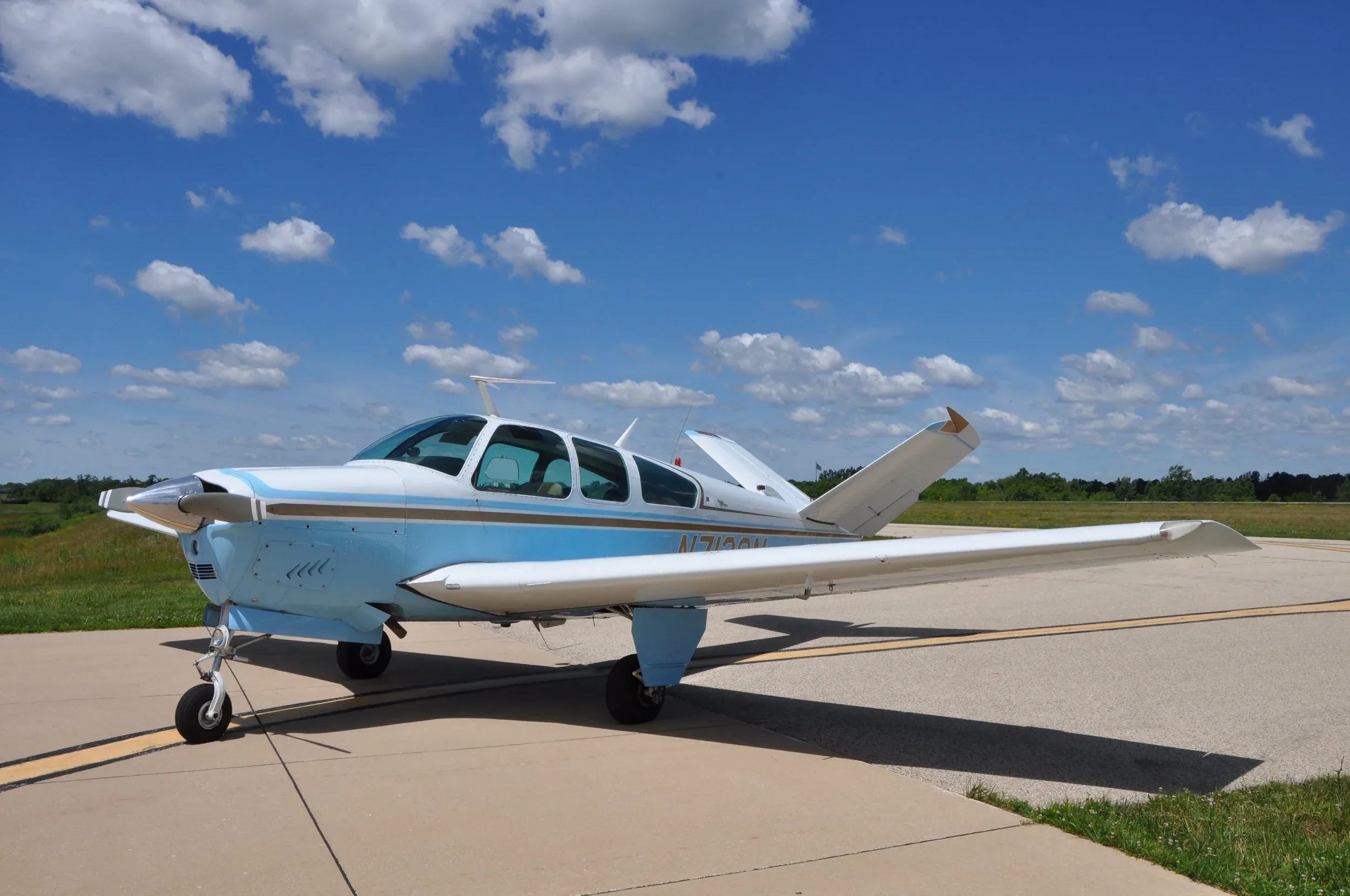 A light blue and white single-engine aircraft with a V-tail parked on an asphalt runway under a partly cloudy sky.