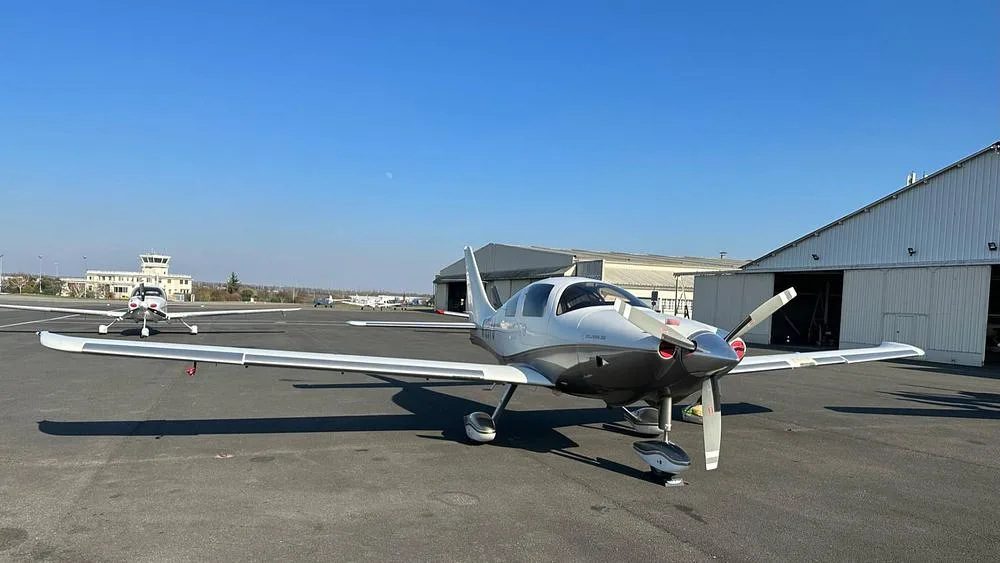 A white and silver Columbia 350 aircraft, N1267J, parked on an airport tarmac with other planes and a control tower in the background under a clear blue sky.