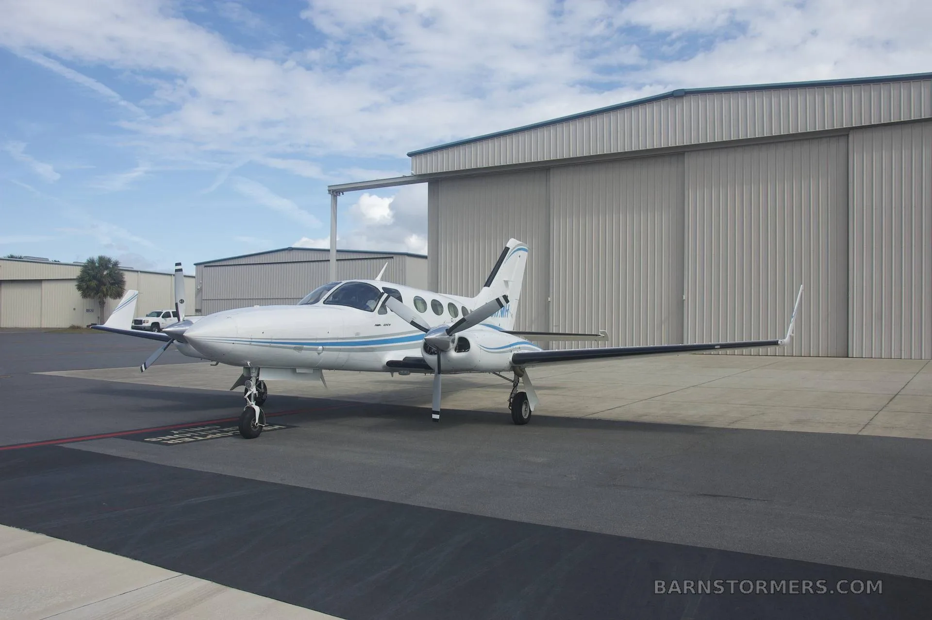 A white twin-engine propeller aircraft with blue and grey stripes (N517MH) is parked on a tarmac with hangars in the background under a partly cloudy sky.